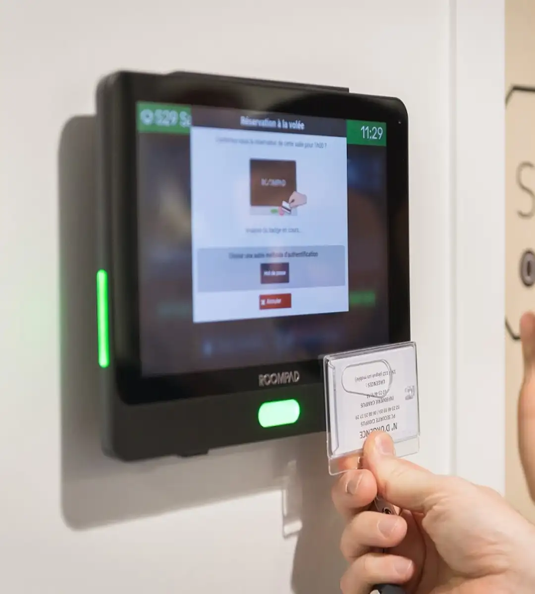 Une main tenant une carte d'identification devant un lecteur de badge électronique RoomPad affichant une interface de réservation à la volée.