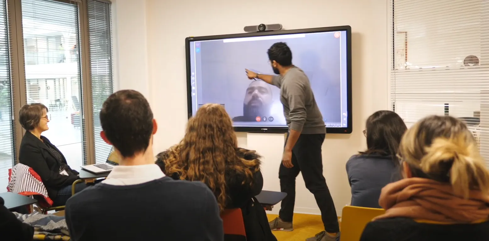 Un homme debout montrant vers un grand écran tactile où un autre homme est en visioconférence, tandis que plusieurs personnes assises regardent la présentation en salle de réunion.