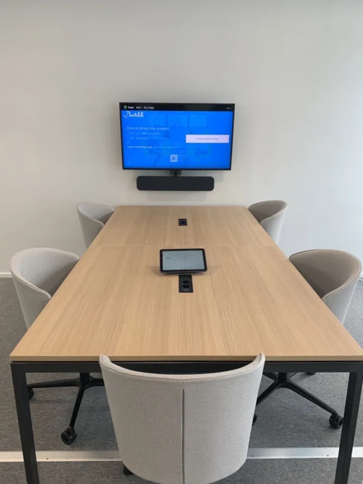 Salle de réunion avec une grande table en bois, cinq chaises grises et un écran mural affichant des instructions de partage d'écran.