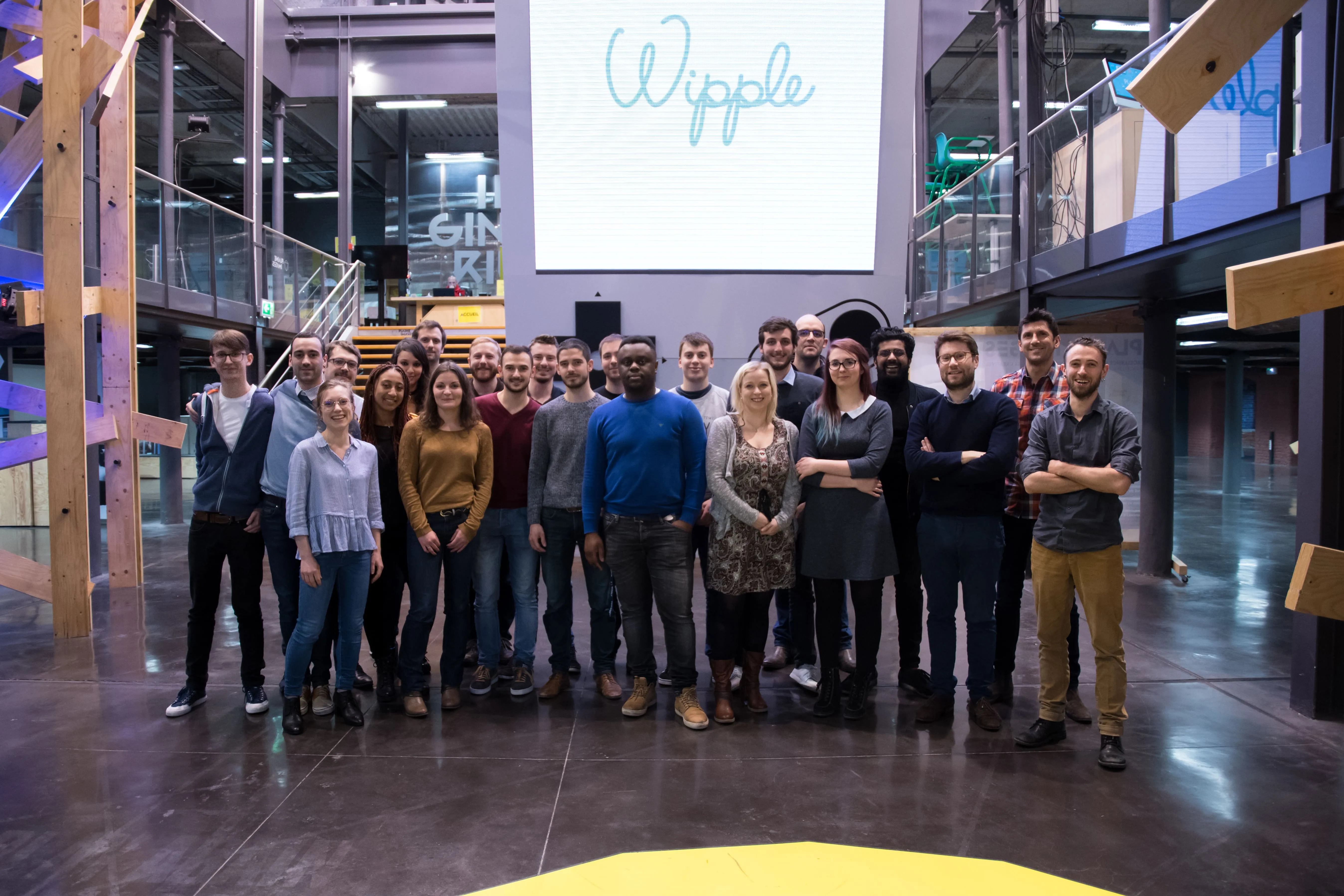 Groupe de vingt-cinq personnes debout dans un grand espace intérieur moderne, avec un grand écran affichant le mot 'Wipple' en arrière-plan.