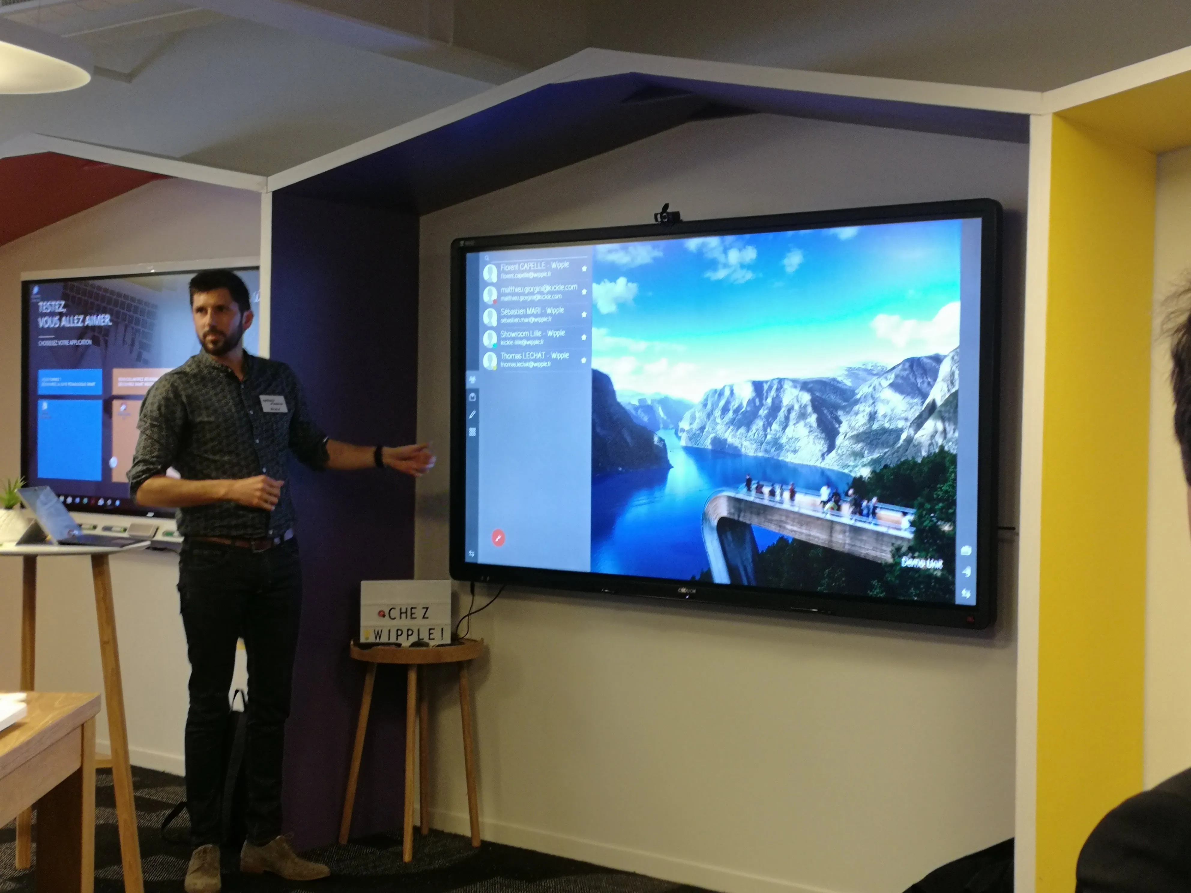 Un homme présente devant un grand écran mural affichant une image de paysage montagnard avec un fjord et un point d'observation.