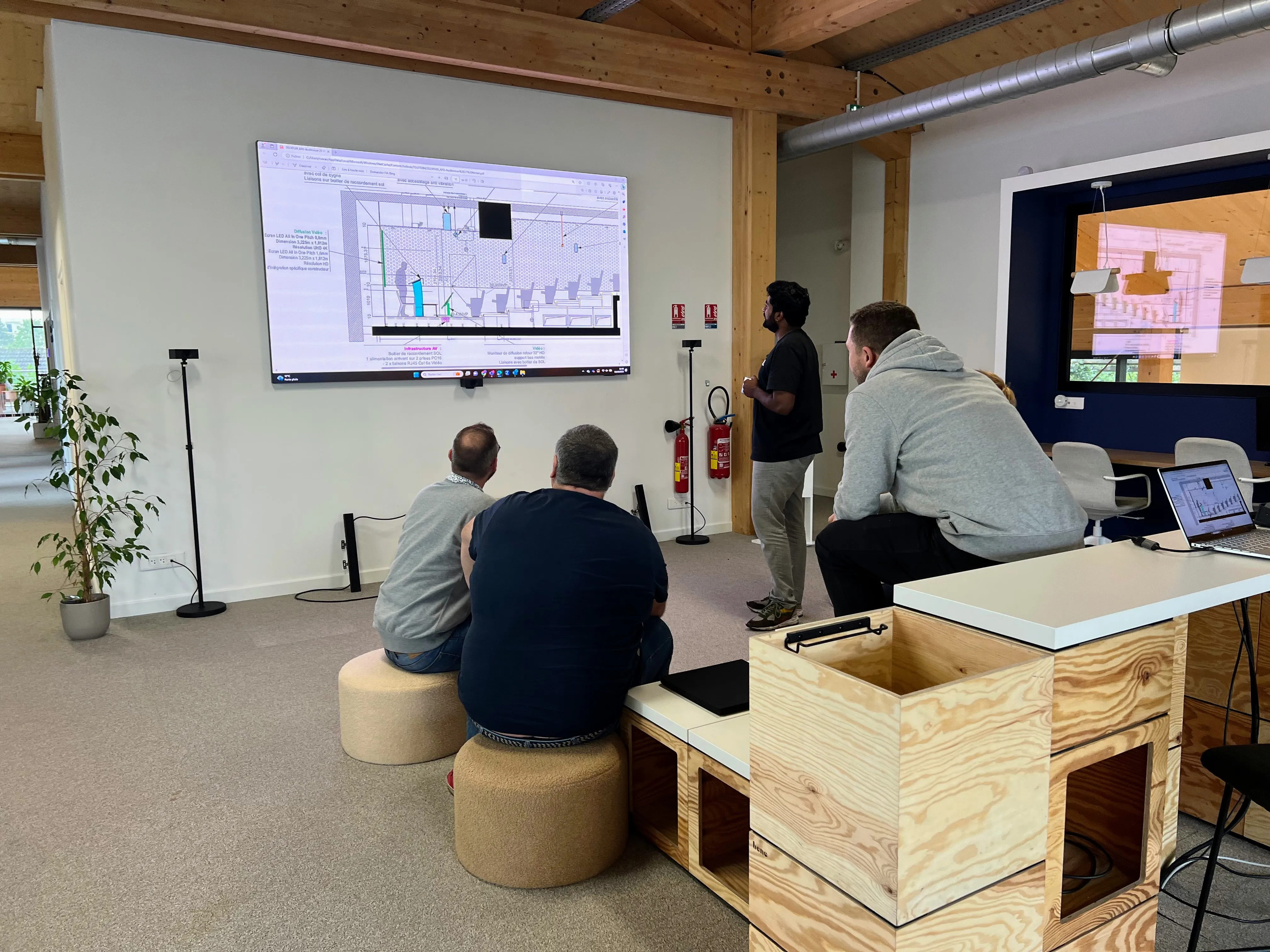 Quatre personnes regardant un écran mural affichant un plan technique dans un bureau moderne avec mobilier en bois clair.