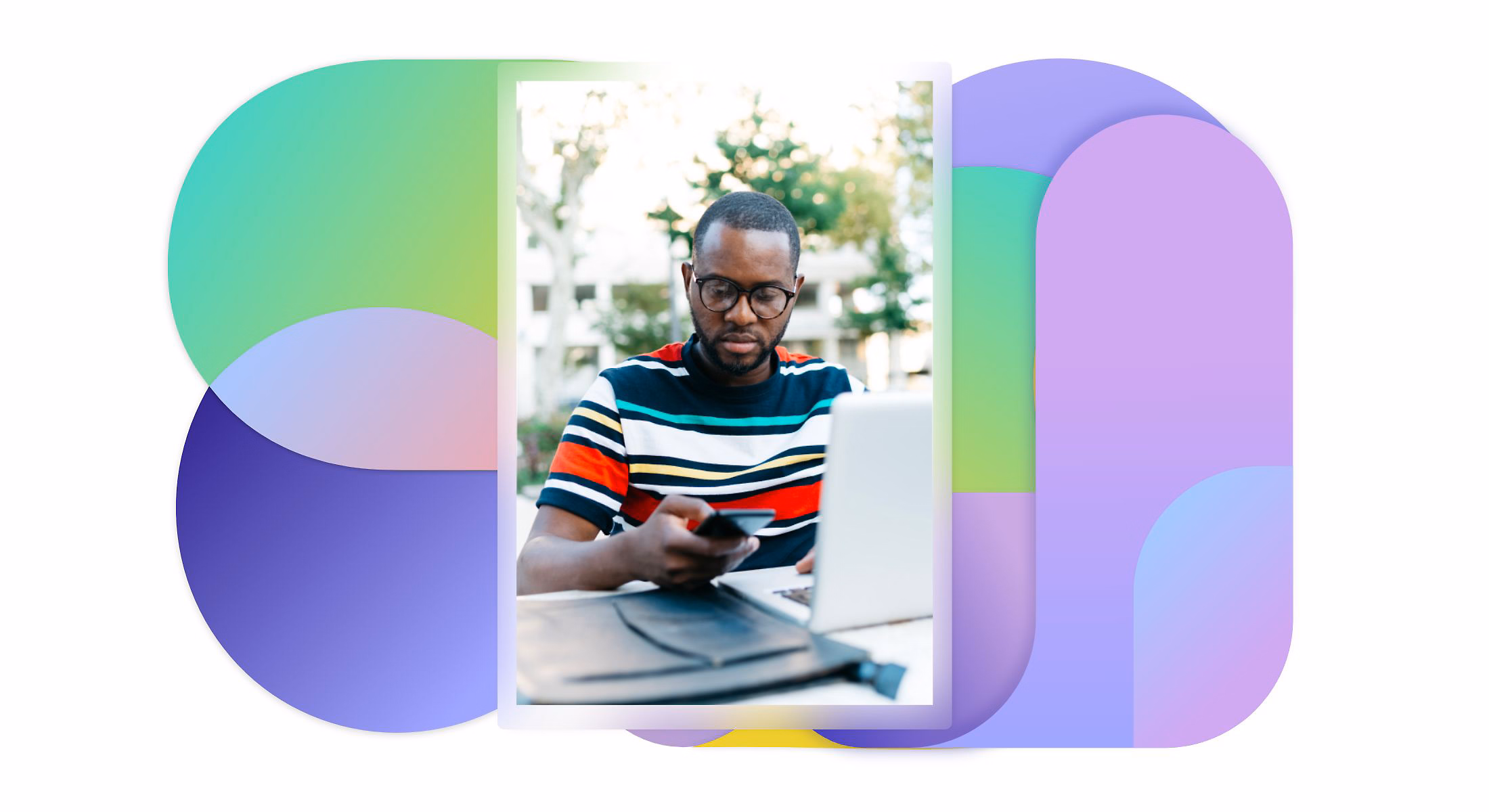 Homme portant des lunettes travaillant sur un ordinateur portable en tenant un smartphone en extérieur.