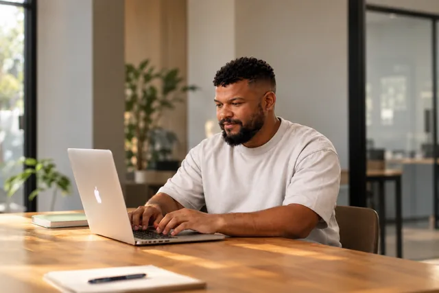 Man typing on a laptop at a wooden table in a sunlit office