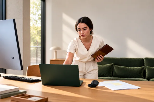 Woman standing at a desk with a laptop, holding a leather folio, in a bright office