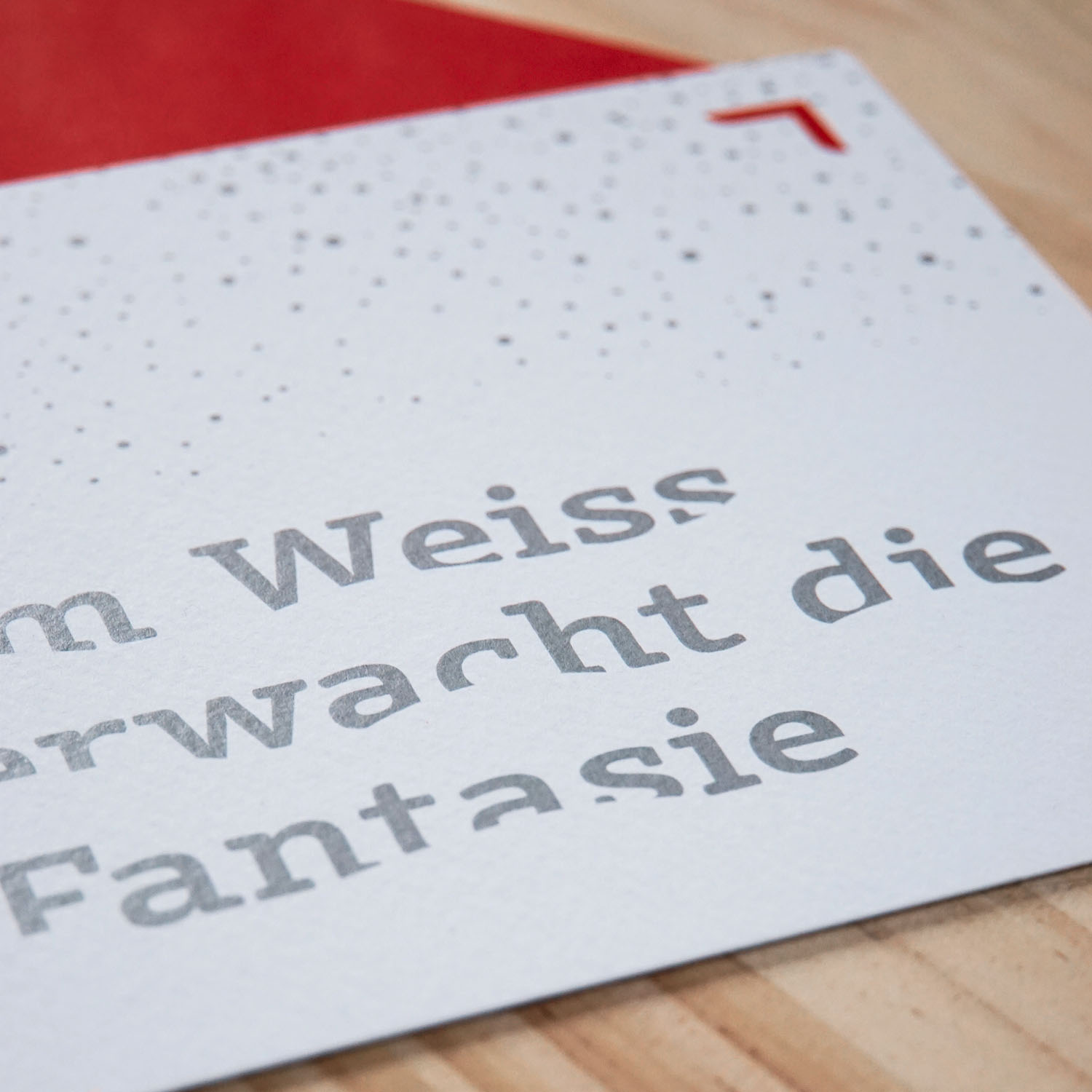 Minimalistische Weihnachtskarte mit silberner Typografie „Im Weiss erwacht die Fantasie“ auf strukturiertem Papier, liegend auf Holz neben rotem Couvert.