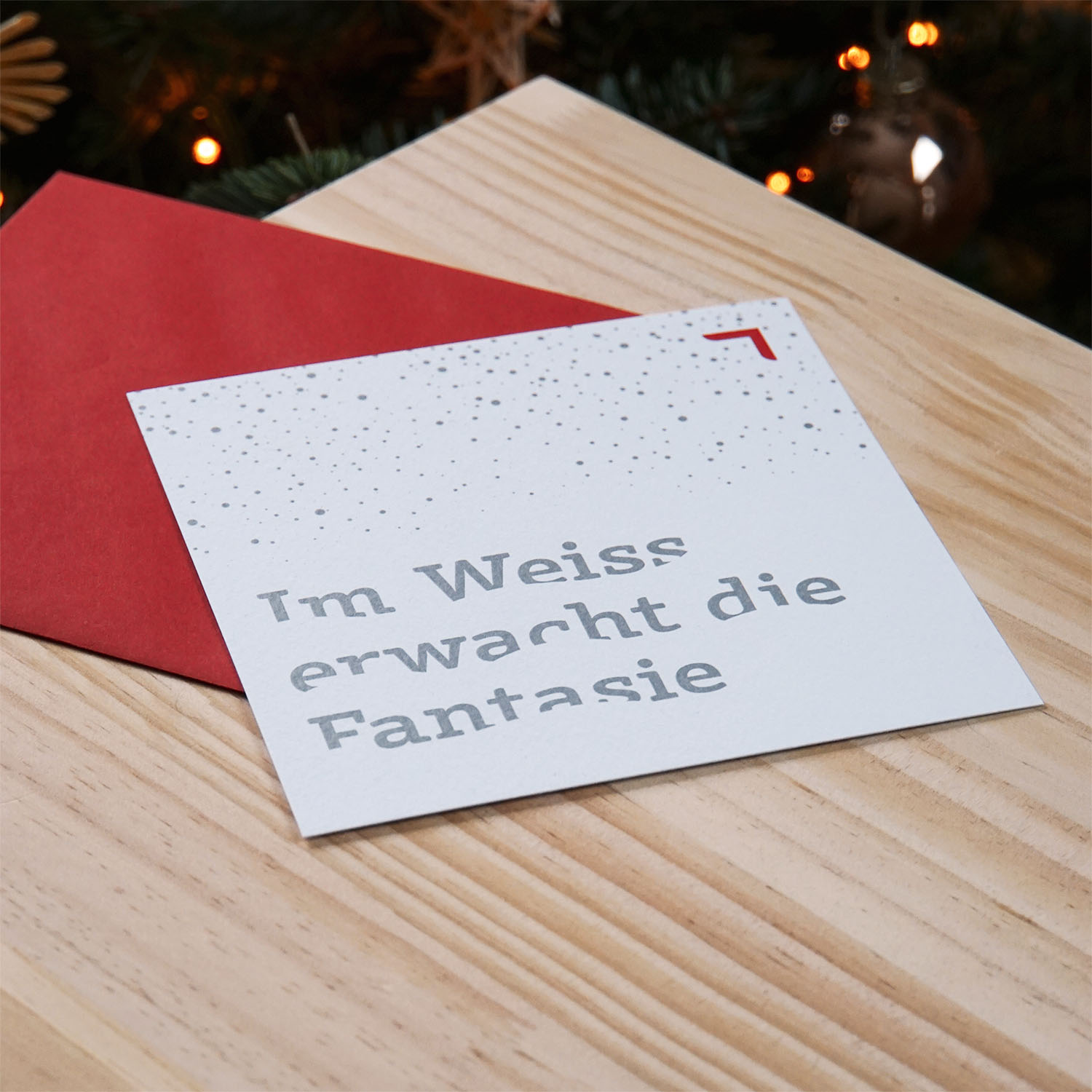 Minimalistische Weihnachtskarte mit silberner Typografie „Im Weiss erwacht die Fantasie“ auf strukturiertem Papier, liegend auf Holz neben rotem Couvert.