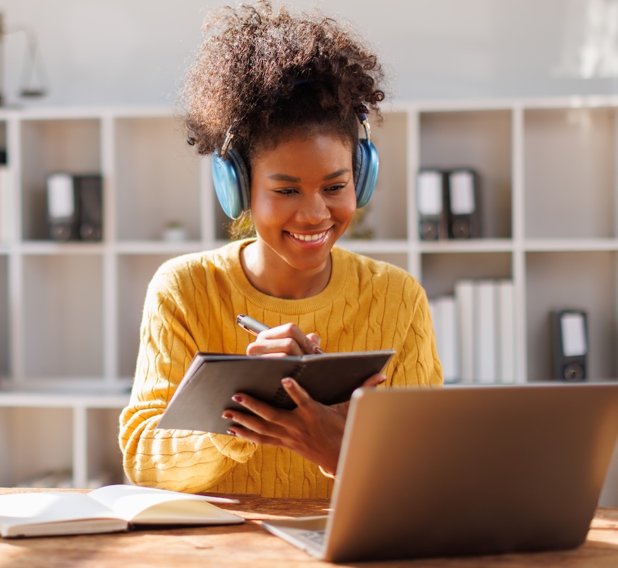 Smiling woman wearing blue headphones, writing in a notebook while looking at a laptop in a bright room.