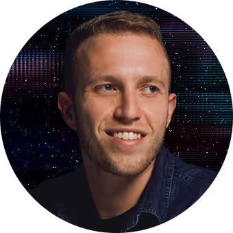 Young man with short hair wearing a denim jacket smiling and looking to the side against a dark background with colorful light patterns.