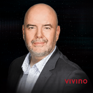 Smiling man with short hair wearing a dark purple checkered shirt against a dark background.