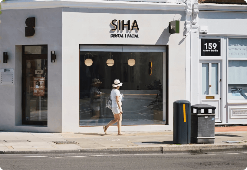 Woman in white hat and dress walking past a storefront with Siha Dental | Facial signage.