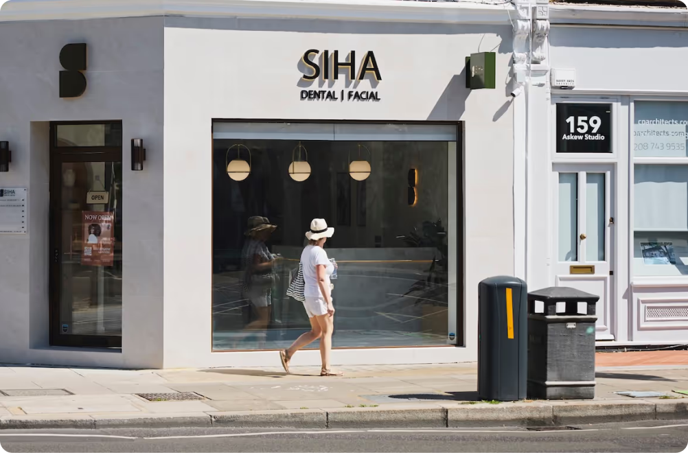 Woman in white hat and dress walking past a storefront with Siha Dental | Facial signage.
