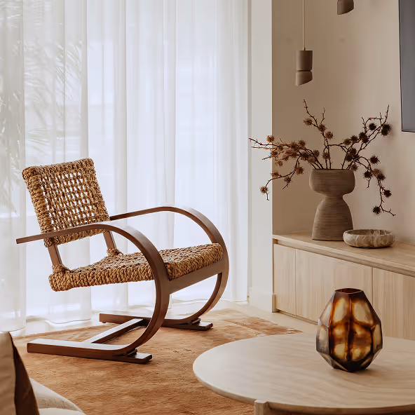 Woven rattan rocking chair in a sunlit living room with beige curtains, a round wooden table with a decorative vase, and a wooden cabinet holding a sculptural vase with dried branches.