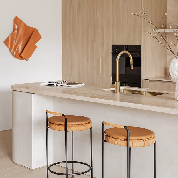 Modern kitchen with curved beige countertop, two leather-topped bar stools, built-in oven, brass faucet, and abstract wall decoration.