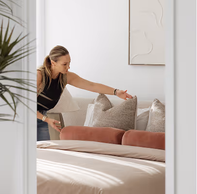 Modern bedroom with a beige upholstered bed, neutral pillows, matching bedside lamps, wooden nightstand, and ocean framed photo on wall.