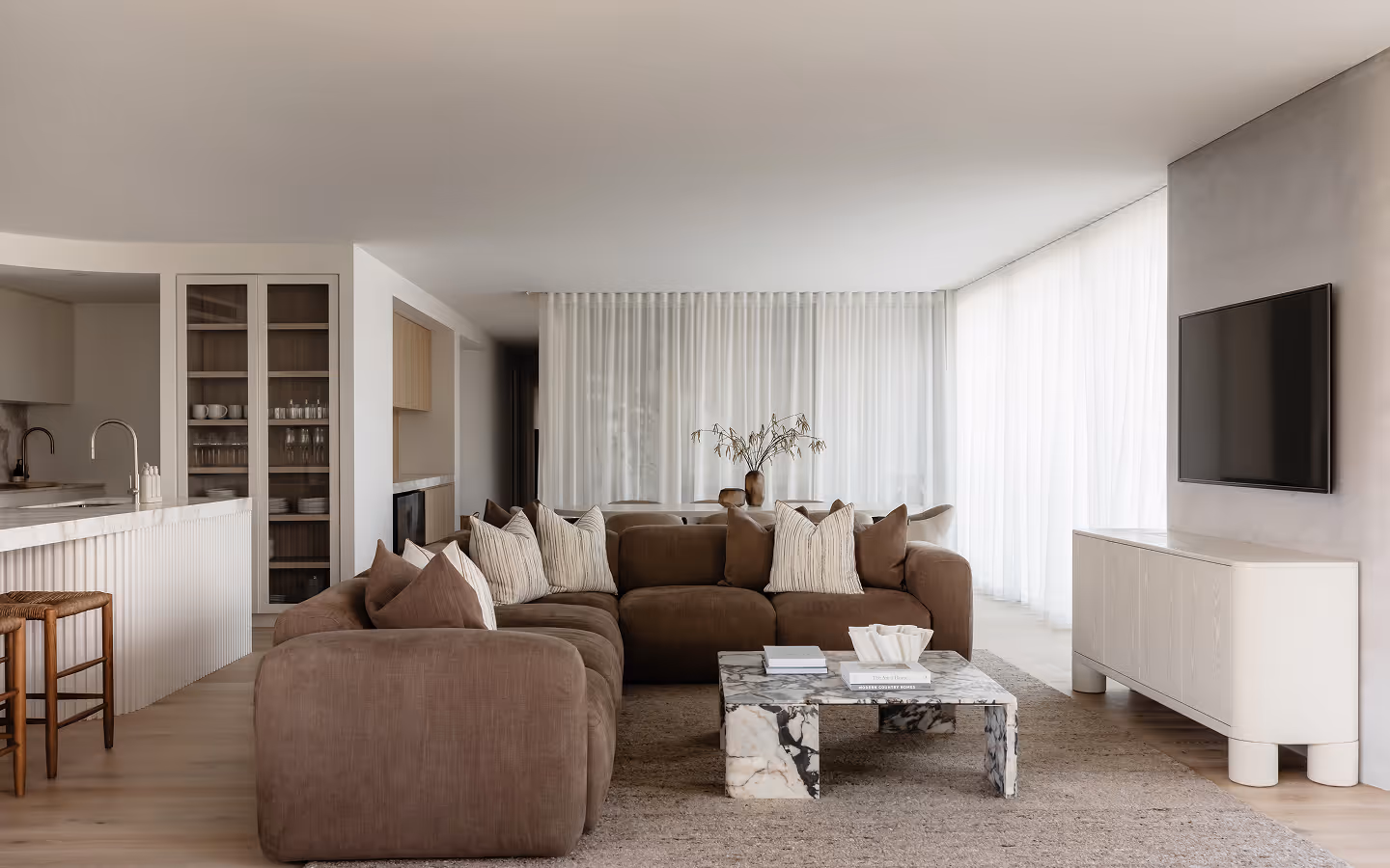 Modern living room with brown sectional sofa, marble coffee table, TV, and white curtains.