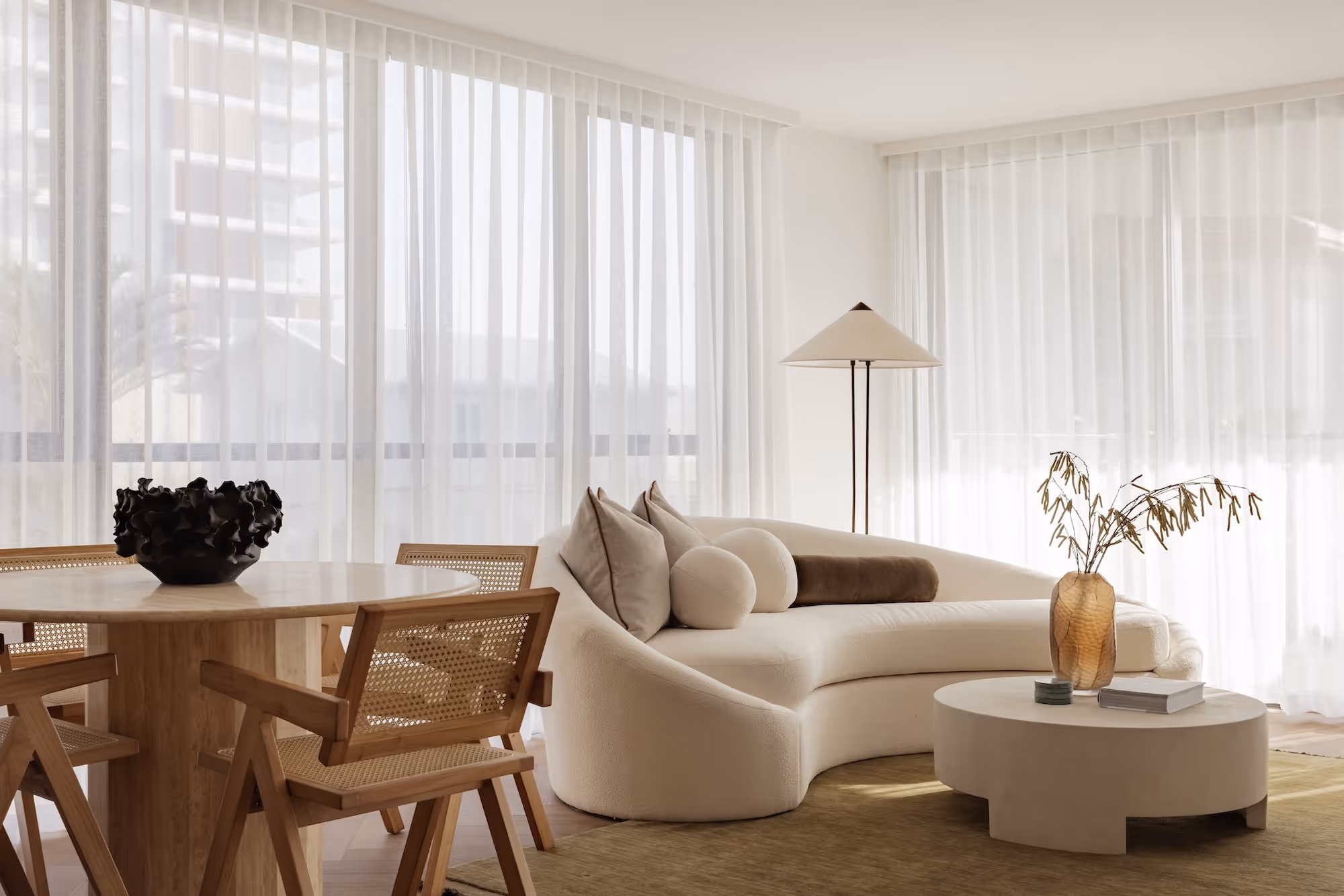 Modern living room with curved white sofa, wood dining table, cane chairs, and minimal decor.