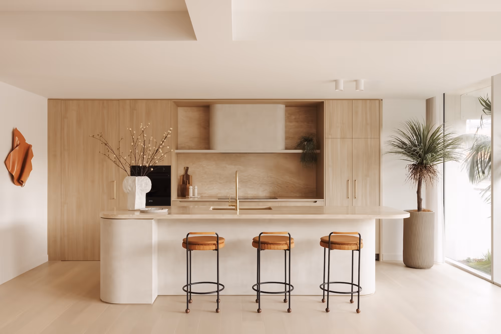 Minimalist kitchen with wood cabinetry, beige island, three stools, a vase, and a tall potted plant.