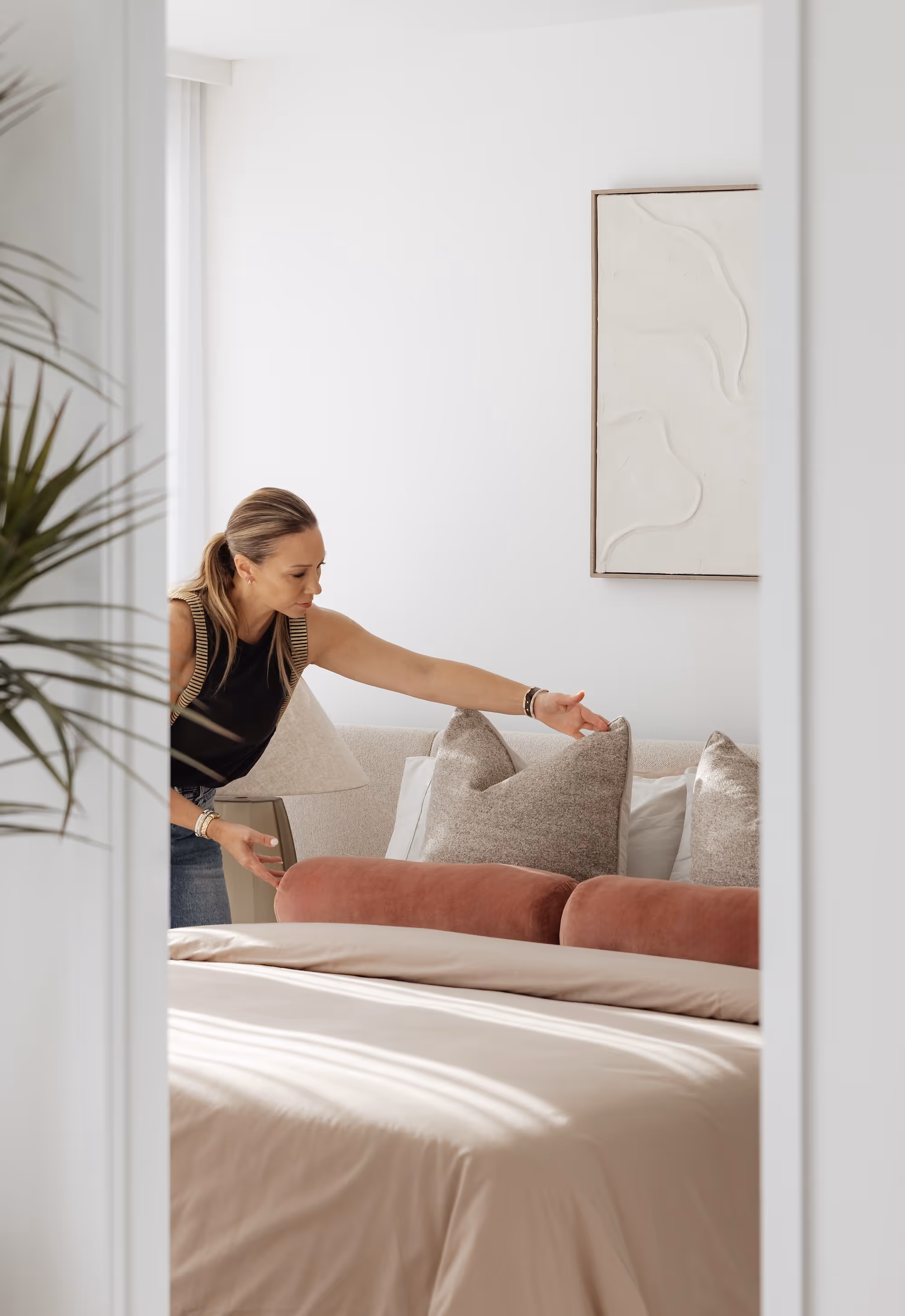 Woman adjusting pillows on a bed in a bright, neutral-toned bedroom with modern decor.