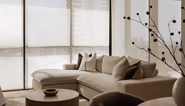 Beige sectional sofa with assorted pillows next to round coffee tables in a sunlit living room.