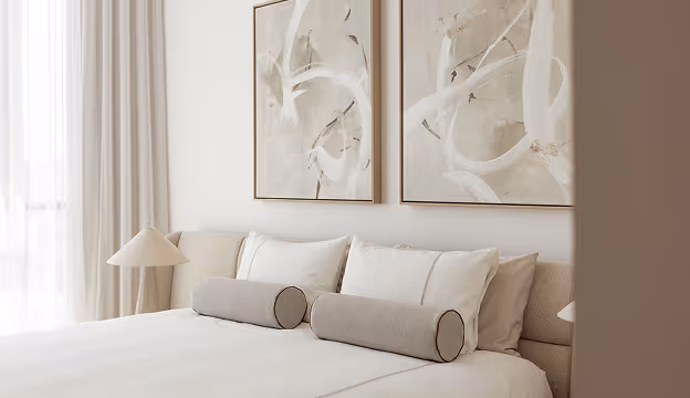 Minimalist bedroom with beige bed, two cylindrical pillows, and abstract art above the headboard.