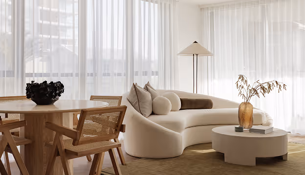 Minimalist room with curved white sofa, wooden round table, wicker chairs, and floor lamp by sheer curtains.