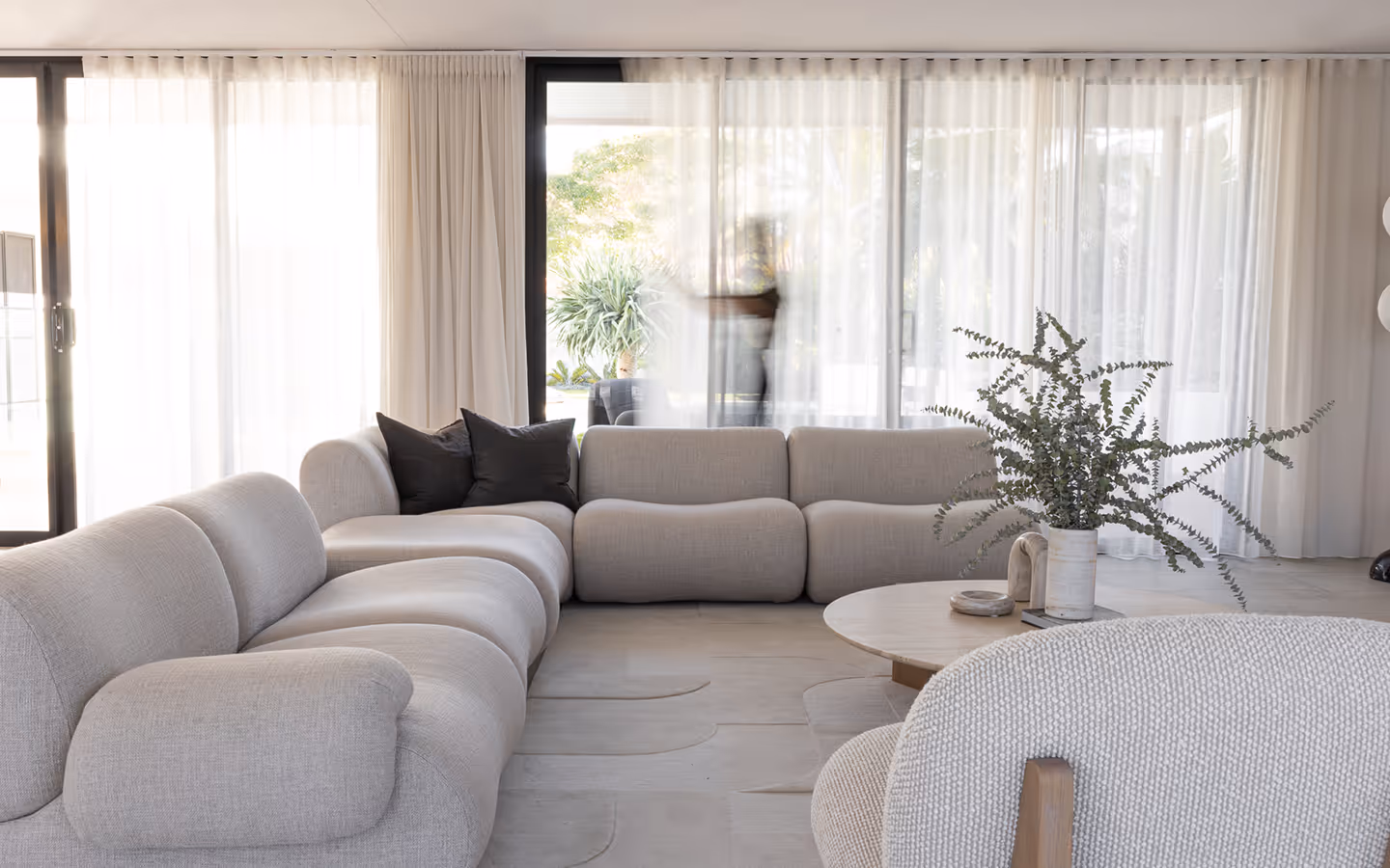 Modern living room with beige sectional sofa, round coffee table, and large sheer curtains covering glass doors.