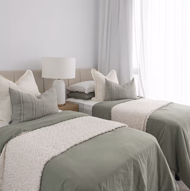 Two twin beds with green bedding, white fluffy runners, pillows, and a lamp between them in a bright room.