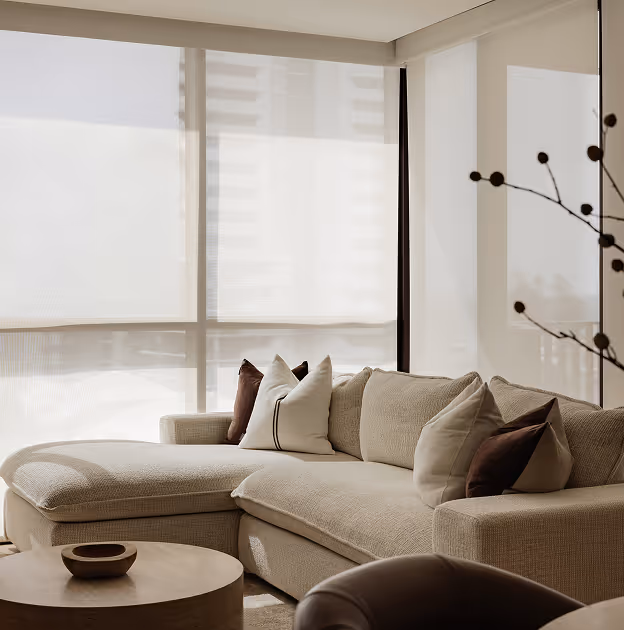 Beige sectional sofa with multiple cushions near large windows covered by translucent roller blinds.
