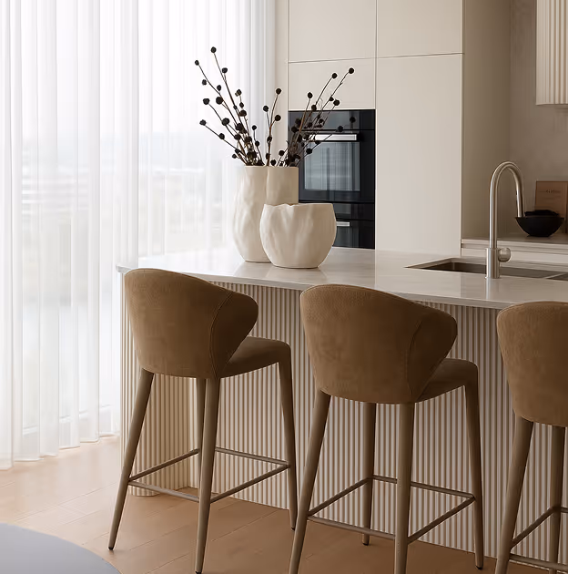 Modern kitchen island with beige bar stools, white countertop, vases with decorative branches, and built-in oven.