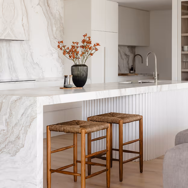 Modern kitchen island with marble countertop, two wooden stools, a vase with orange flowers, and a silver faucet.