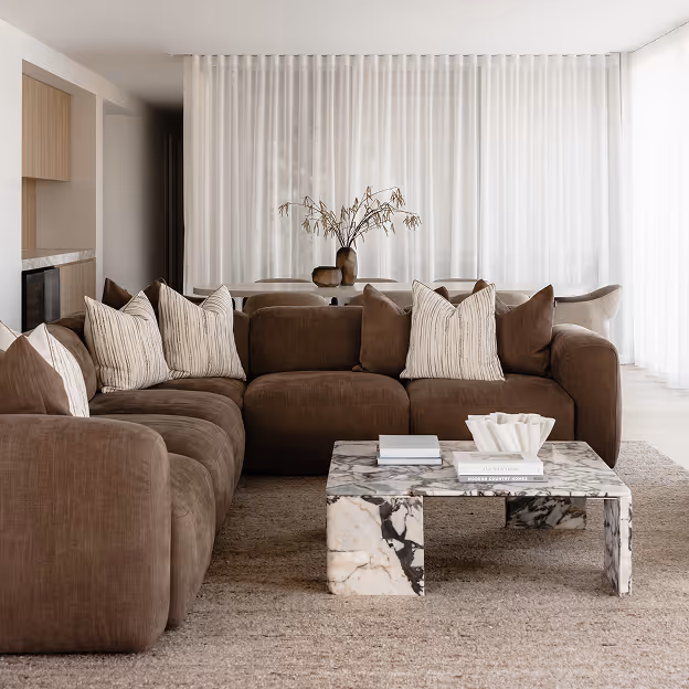 Living room with brown sectional sofa, patterned pillows, marble coffee table, and sheer white curtains.