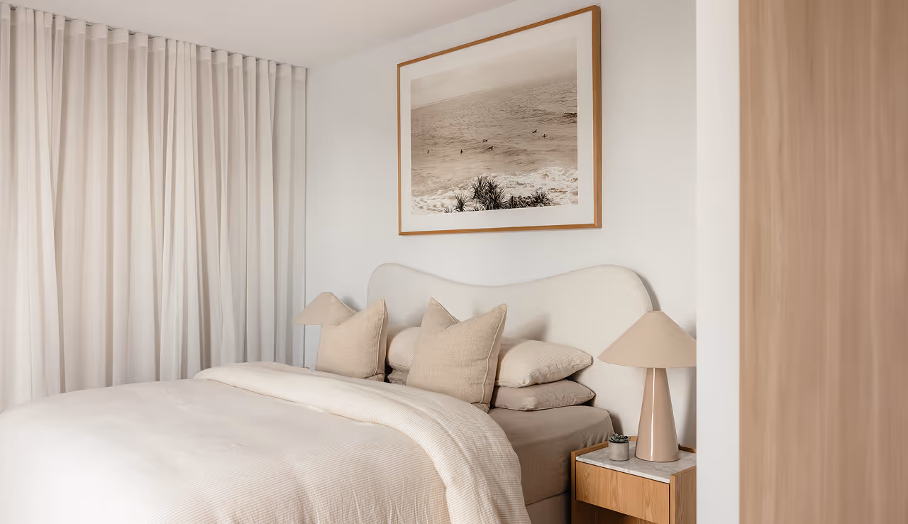 Minimalist bedroom with beige bedding, pillows, lamps, a wooden nightstand, and ocean photo on wall.