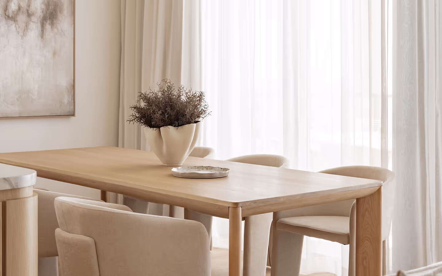 Minimalist wooden dining table with beige chairs, vase of dried branches, and sheer white curtains.