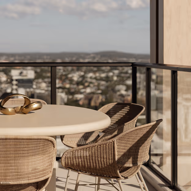 Round table with decorative rings and four wicker chairs on a balcony with city view and glass railing.