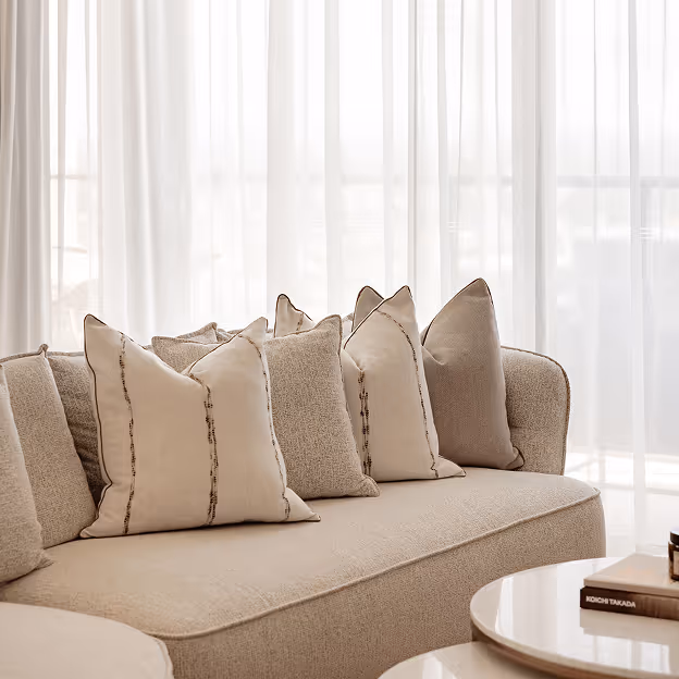 Beige curved sofa with several light-color textured throw pillows in front of sheer white curtains.