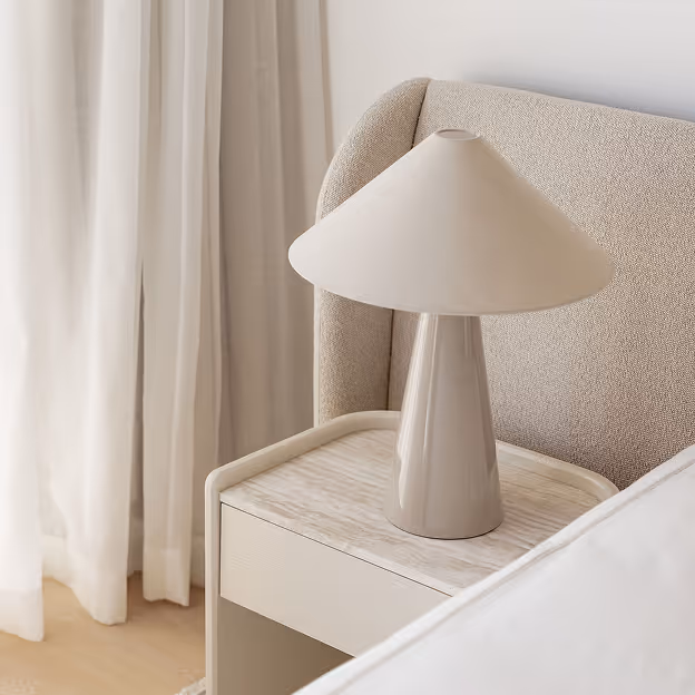 Beige table lamp with a conical shade on a light wooden bedside table next to a textured beige headboard.