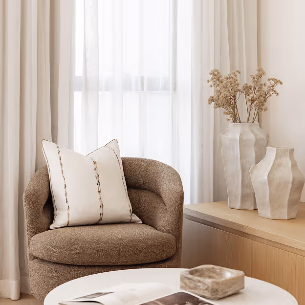 Cozy brown armchair with white pillow beside wooden table holding two cream vases and dried flowers.