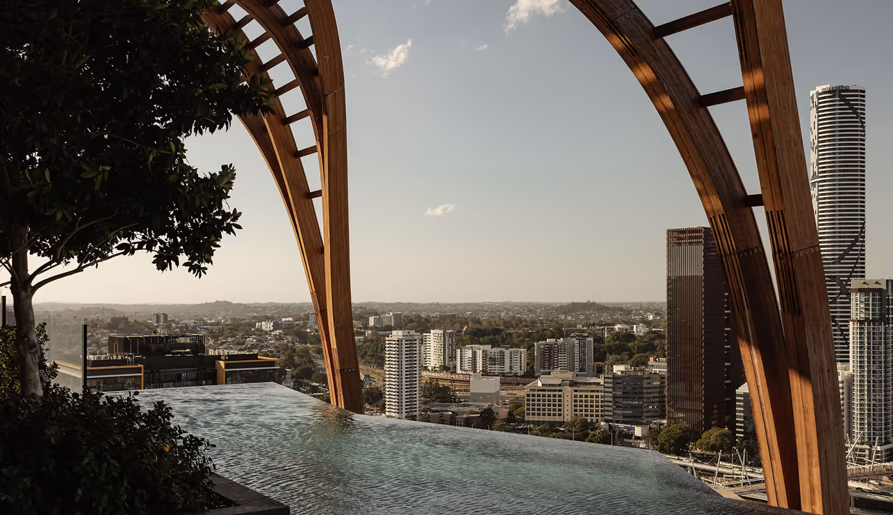 Infinity pool with wooden arches overlooks urban cityscape under clear sky at sunset.