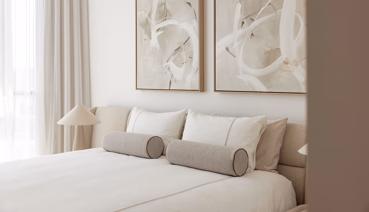 Minimalist bedroom with beige and white bed linens, cylindrical pillows, and abstract wall art.