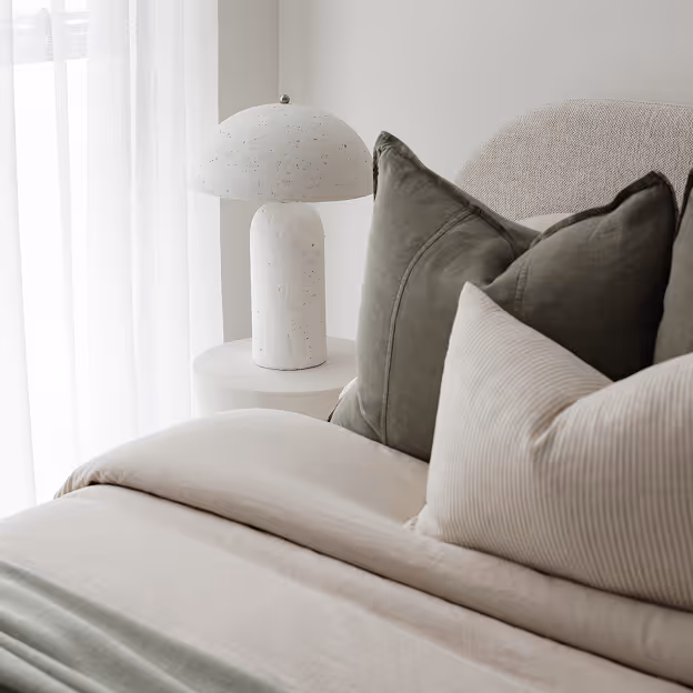 Modern beige lamp with a cone-shaped shade on a light wood nightstand next to a textured headboard.