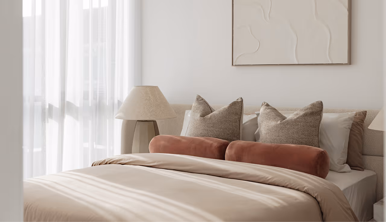 Neutral-toned bedroom with white and beige pillows, bolsters, abstract wall art, and a lamp near sheer curtains.