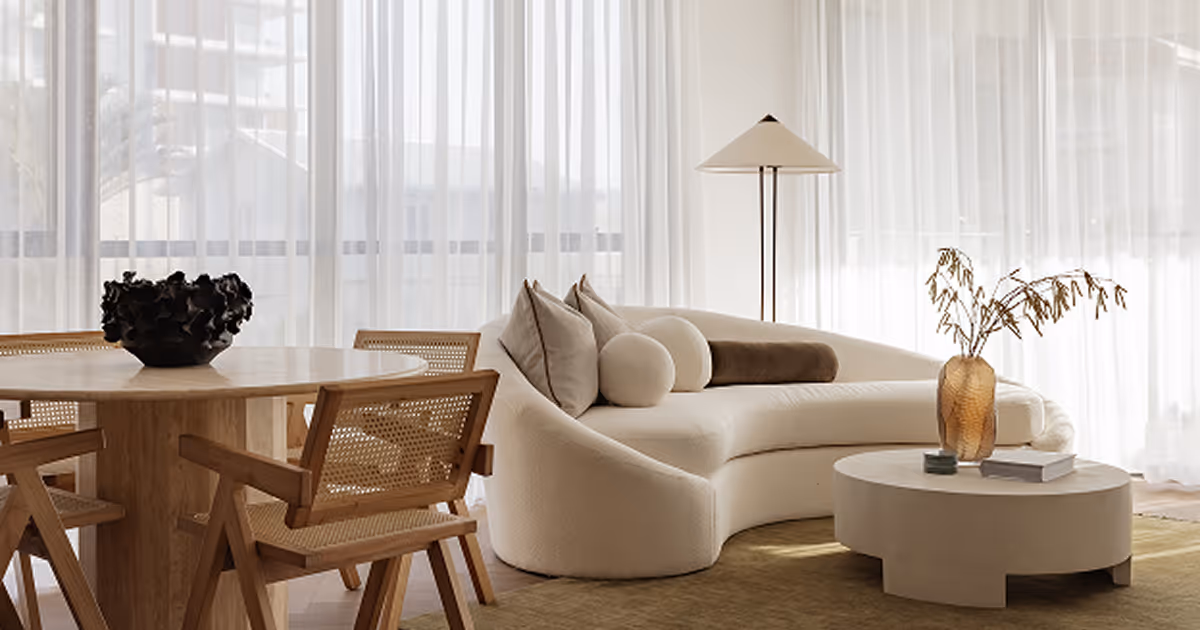 Minimalist living room with a curved beige sofa adorned with various cushions, a round white coffee table with a brown glass vase holding dried plants, a floor lamp with a conical shade, and a wooden dining table with matching cane chairs near large sheer white curtains.