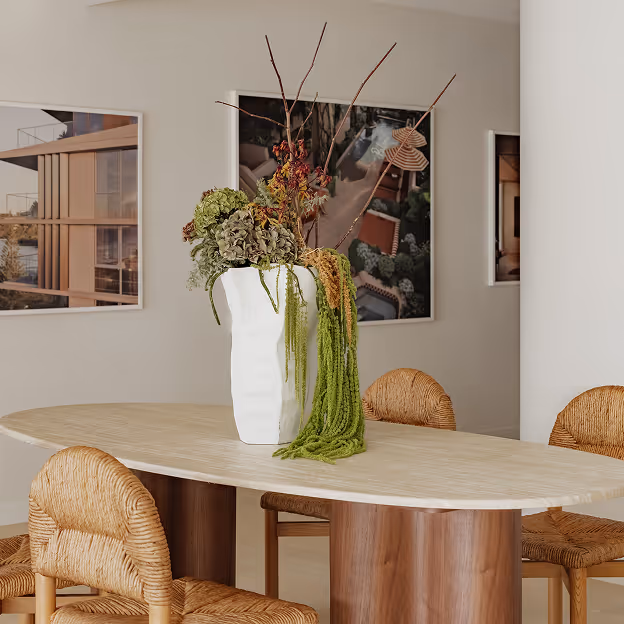 Modern dining table with wooden legs, wicker chairs, and a tall white vase with dried flowers and green strands.