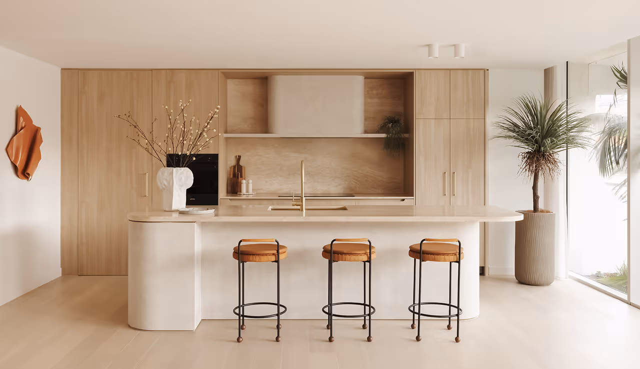 Modern kitchen with wooden cabinetry, white island, three stools, tall plant, and decorative vase with branches.