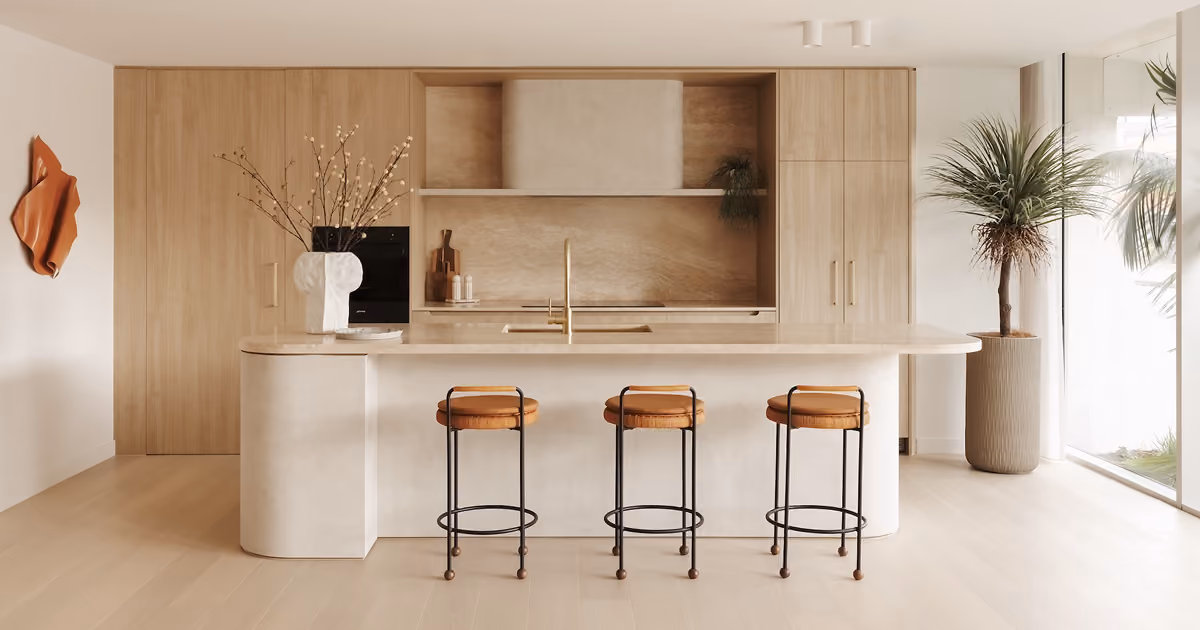 Minimalist kitchen with light wood cabinetry and a large beige marble island with a gold faucet, three bar stools with brown leather seats, a white abstract vase with branches on the island, and a tall potted plant by a large window on the right.