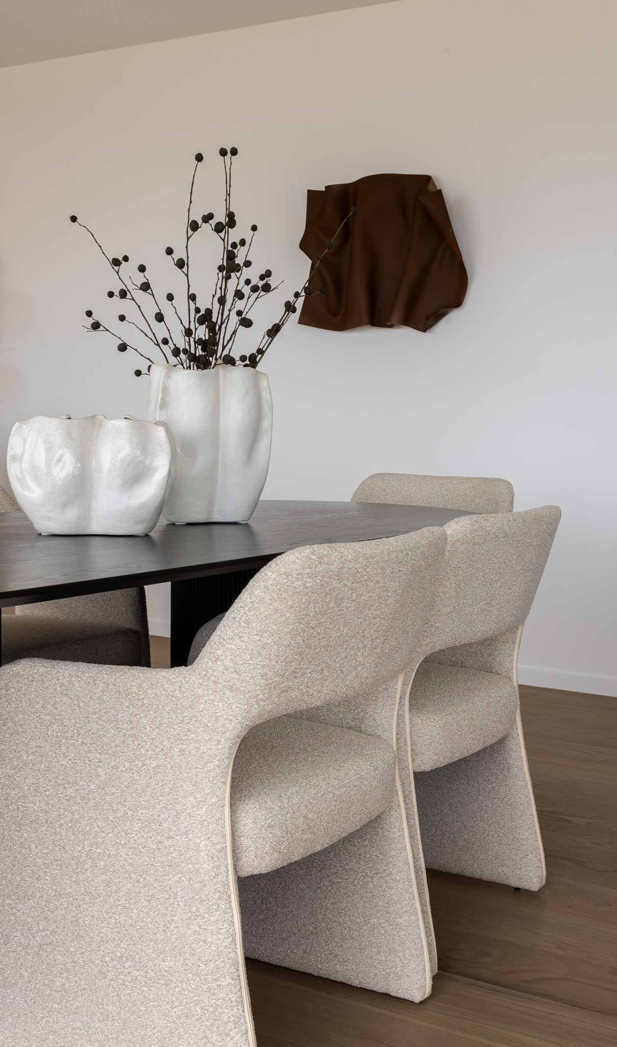 Modern dining table with wooden legs, wicker chairs, and a tall white vase with dried flowers and green strands.