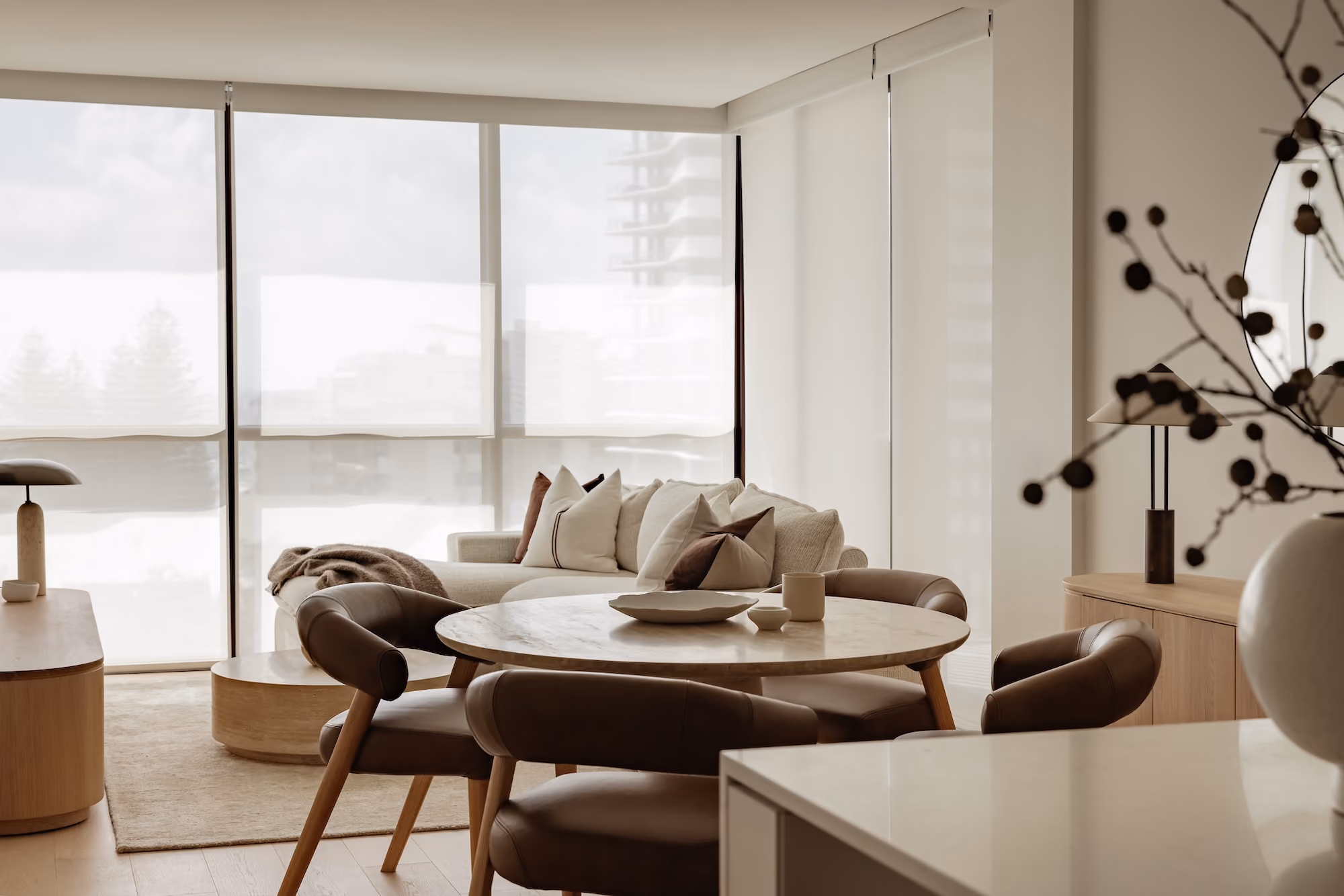 Light wood dining table with modern cream chairs and ceramic vase with dried branches near sheer white curtains.