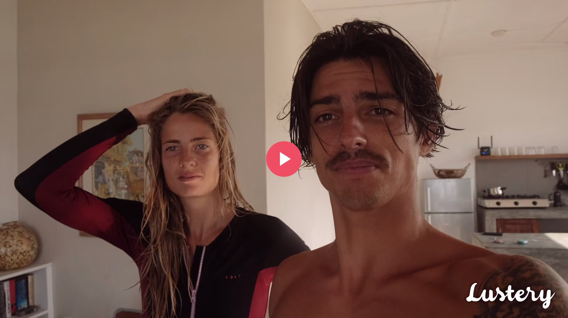 James and Lola smile at the camera in wetsuits in their home after surfing.