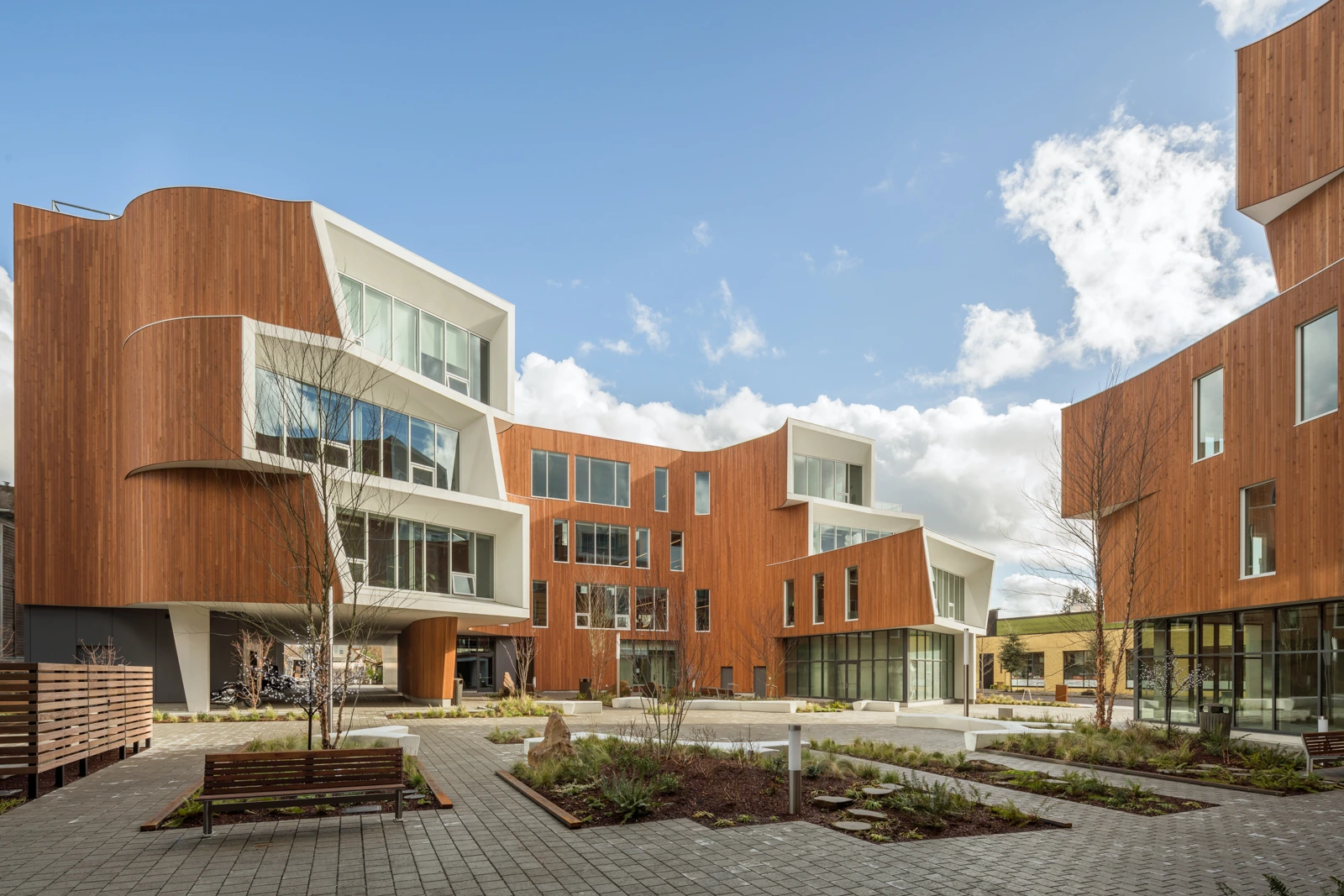 Completed mixed‑use complex with wood‑clad façades and white cantilevered apertures, surrounded by landscaped courtyards and walkways.